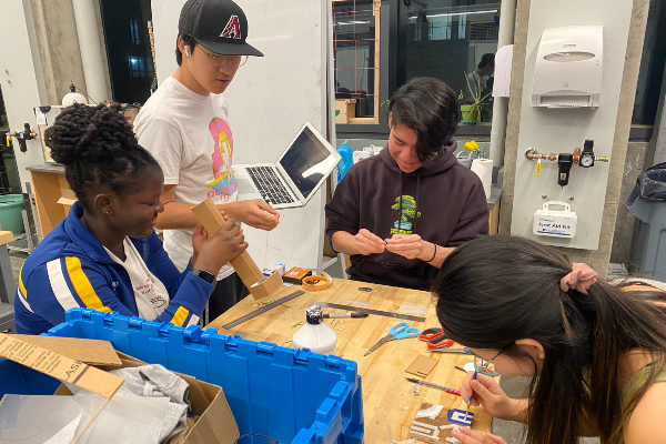 Three students sitting around table working on prototype development with one student standing with laptop