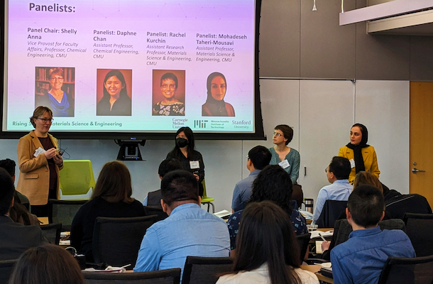 Four panelists standing in front of presentation screens talking to seated participants