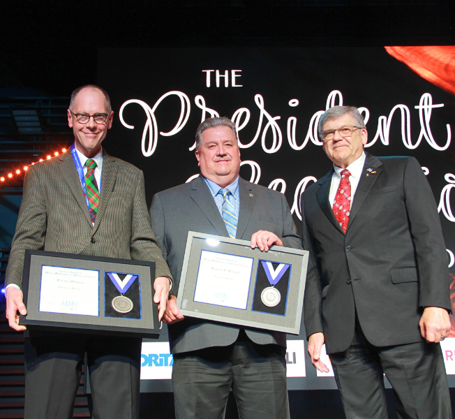 Three men standing, two holding awards
